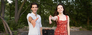A young man and woman in casual summer attire, smiling and offering a handshake in a lush park setting.
