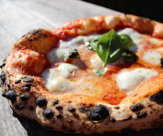 A rustic wood-fired pizza with a charred crust, melted mozzarella, sliced tomatoes, and fresh basil leaves