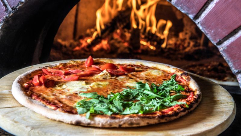A tomato and greens pizza in front of a wood-fired oven with flames.