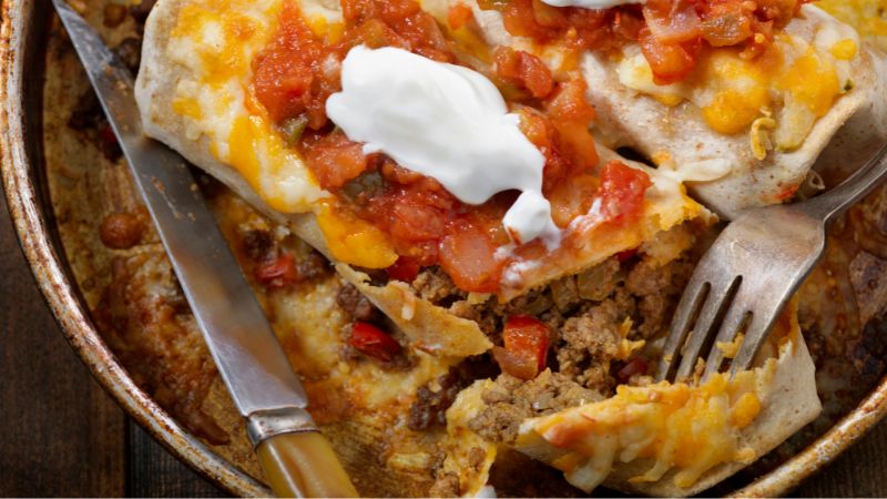 Close-up of a cut enchilada with salsa, melted cheese, sour cream, and a knife and fork on the plate.