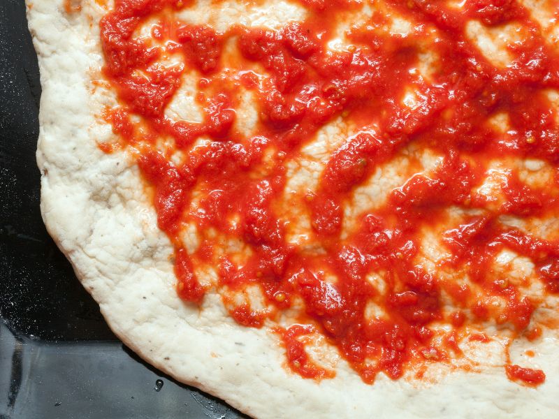 Close-up of pizza dough partially topped with tomato sauce.