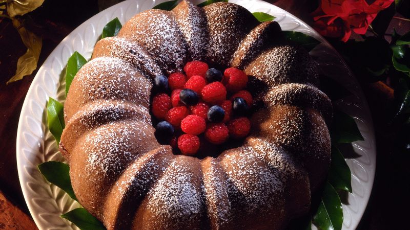 A Bundt cake with powdered sugar, raspberries, and blueberries on a white plate surrounded by leaves.