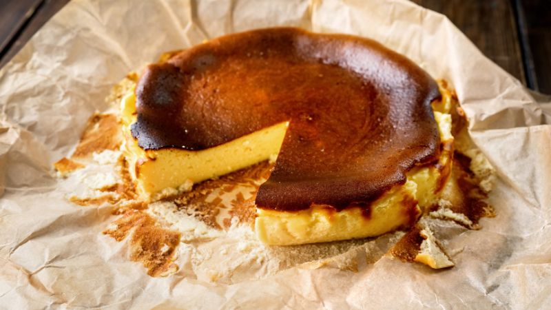 A partly eaten burnt cheesecake on parchment paper shows a creamy middle and dark caramelized top.