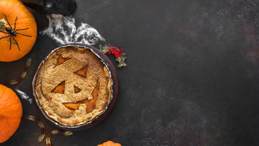 A pumpkin pie with a carved face is surrounded by small pumpkins, plastic spiders, and cobwebs on a dark surface.