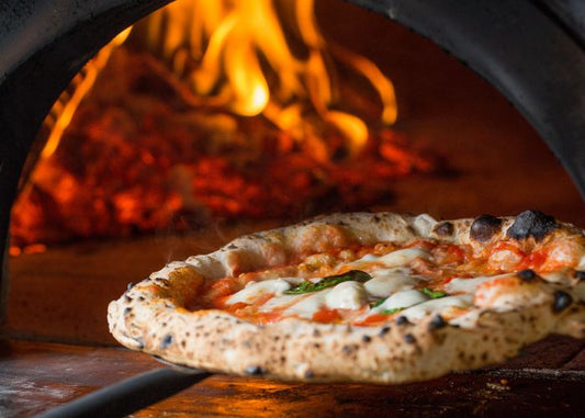 A freshly baked, thin-crust pizza being cooked in a wood-fired oven, with melted cheese, toppings, and the warm orange glow of the oven flames creating an inviting, authentic Italian culinary scene.