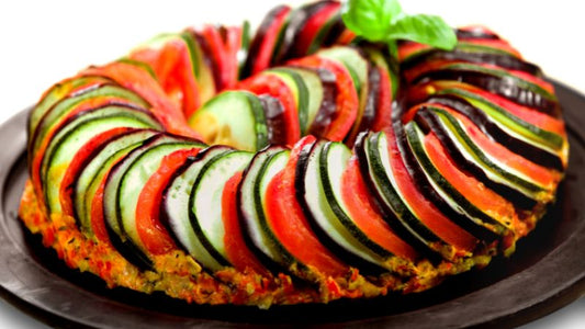 Colorful ratatouille in a circular pattern on a dark plate, topped with a basil leaf.