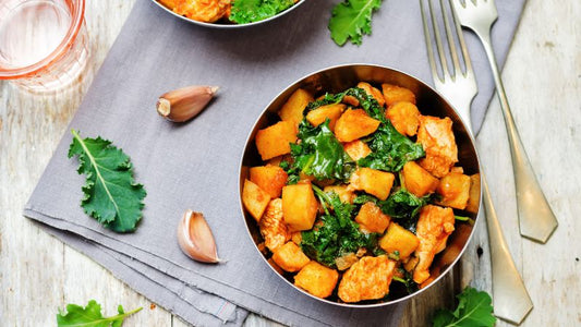 A bowl of roasted sweet potatoes and kale with garlic on a gray napkin, next to a fork and a glass of water.