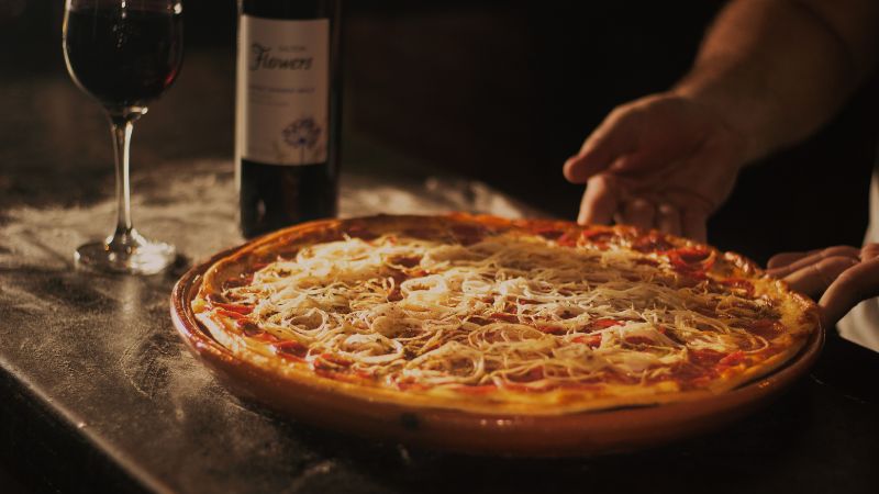 A pizza with cheese and toppings is placed next to a bottle and glass of red wine on a table.