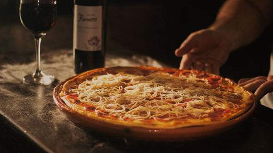 A pizza with cheese and toppings is placed next to a bottle and glass of red wine on a table.