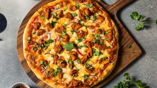 A cooked pizza on a wooden board with chicken, cheese, red onions, and herbs; scattered ingredients and utensils surround it.