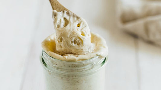 A wooden spoon lifts creamy substance from a glass jar on a light surface.