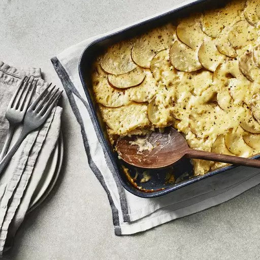 A baking dish of scalloped potatoes on a cloth, with a wooden spoon inside; plates and forks are beside it.