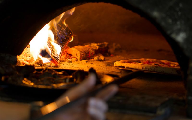 A pizza being slid into a wood-fired oven with visible flames on the side.