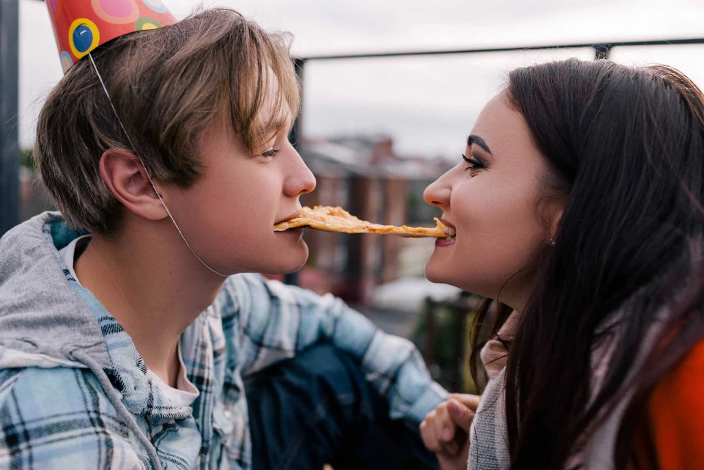 Two people, one in a party hat, share a slice of pizza from opposite ends outside.