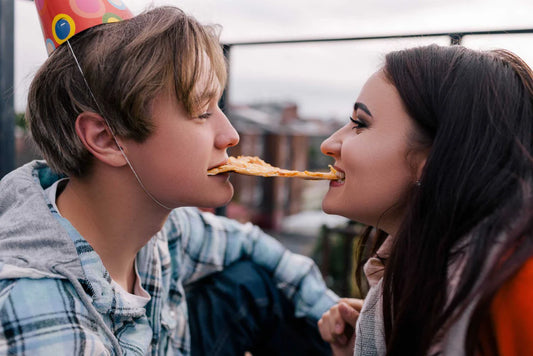 Two people, one in a party hat, share a slice of pizza from opposite ends outside.