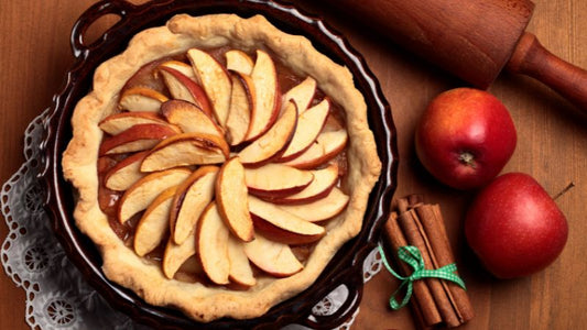 Apple pie topped with slices in a brown dish, with a rolling pin, two red apples, and cinnamon sticks on a wooden table.