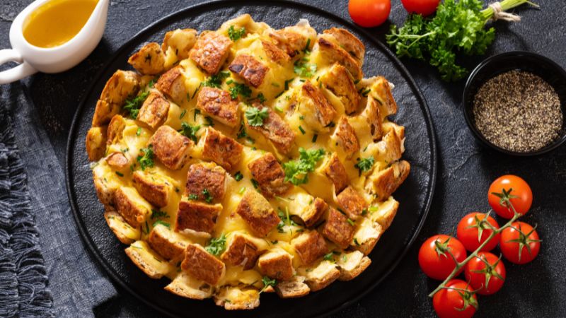 Cheese and herb pull-apart bread on a black plate, with cherry tomatoes, parsley, pepper bowl, and sauce container on a dark surface.
