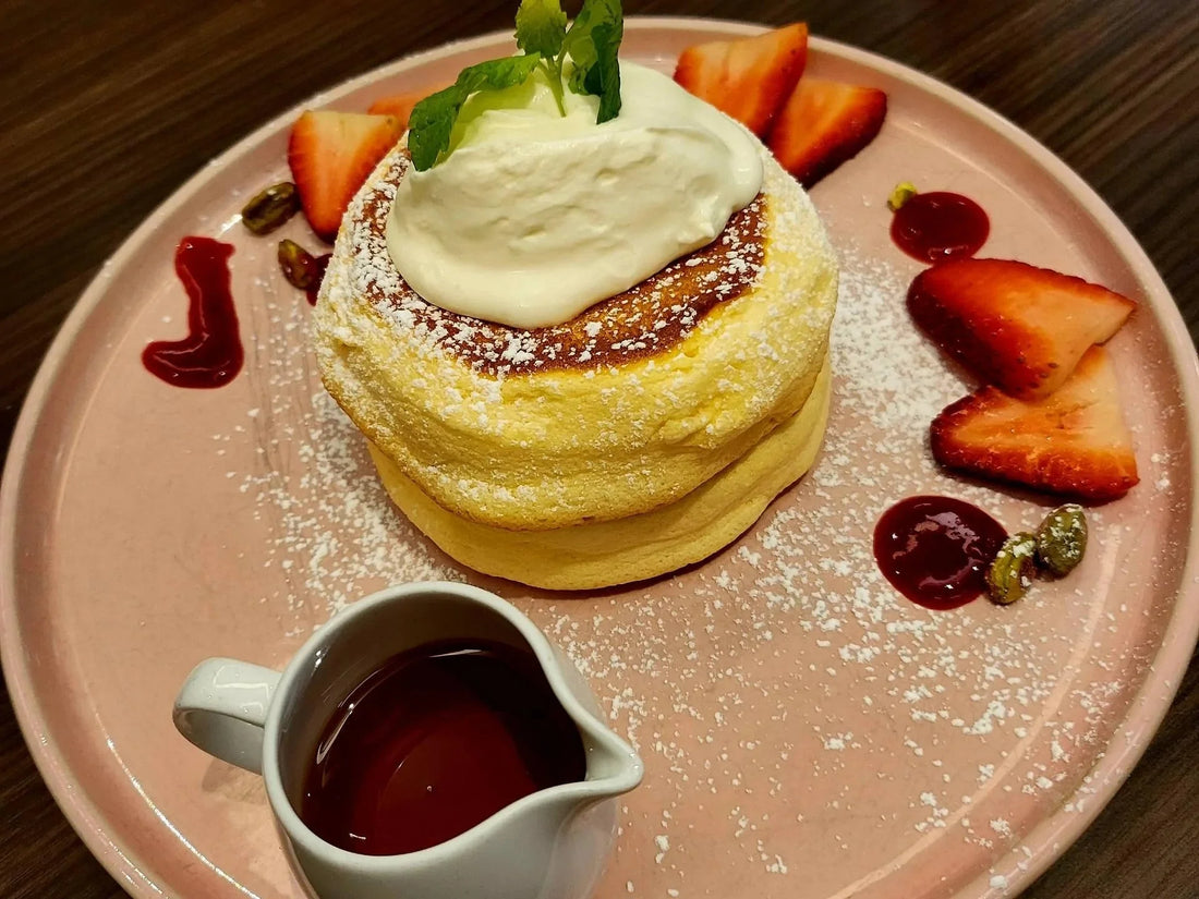 A fluffy pancake topped with whipped cream and a mint leaf, surrounded by strawberries and syrup on a pink plate, with a small syrup pitcher.