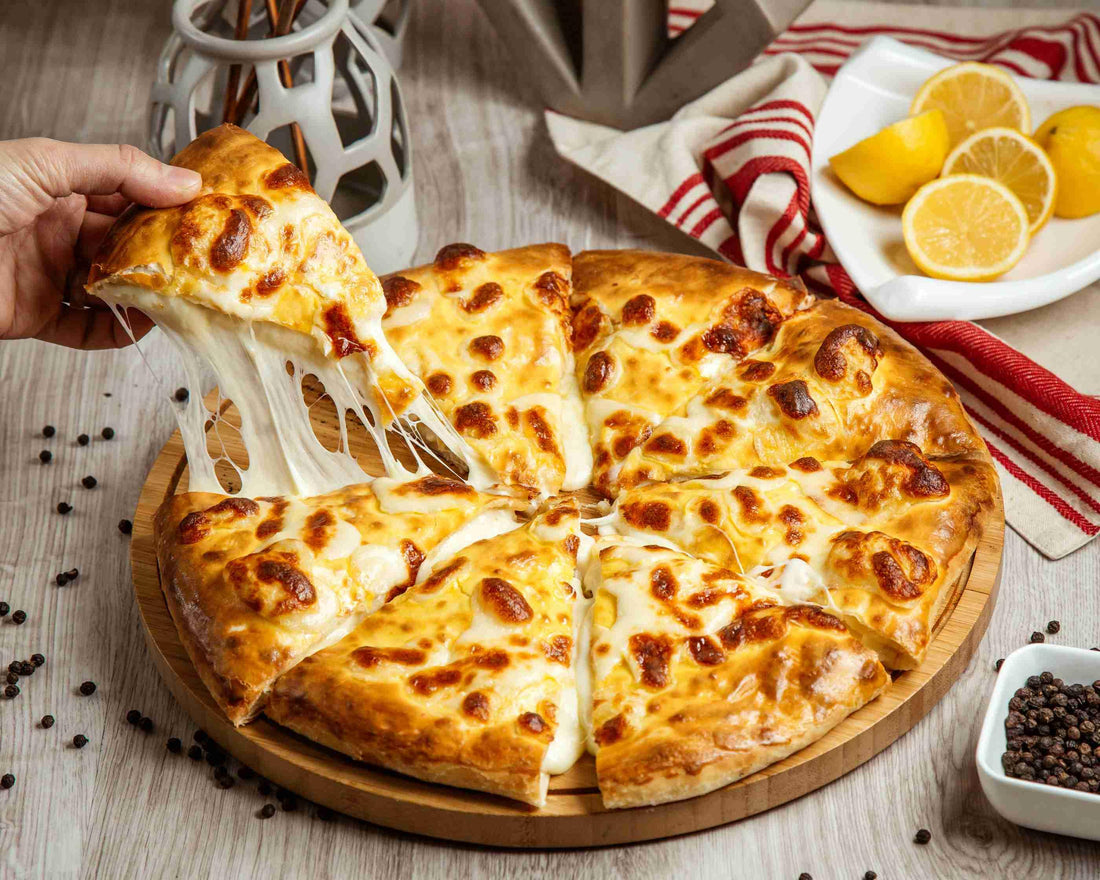 A hand pulls a cheesy pizza slice from a wooden tray next to lemon wedges, a red and white cloth, and scattered peppercorns.
