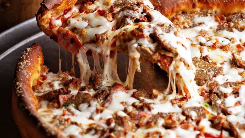 A slice of cheesy pizza with sausage and pepperoni is lifted from a round pan, revealing strands of melted cheese.