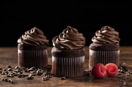 Three chocolate cupcakes with frosting, two raspberries, and scattered chocolate chips on wood. Black background.