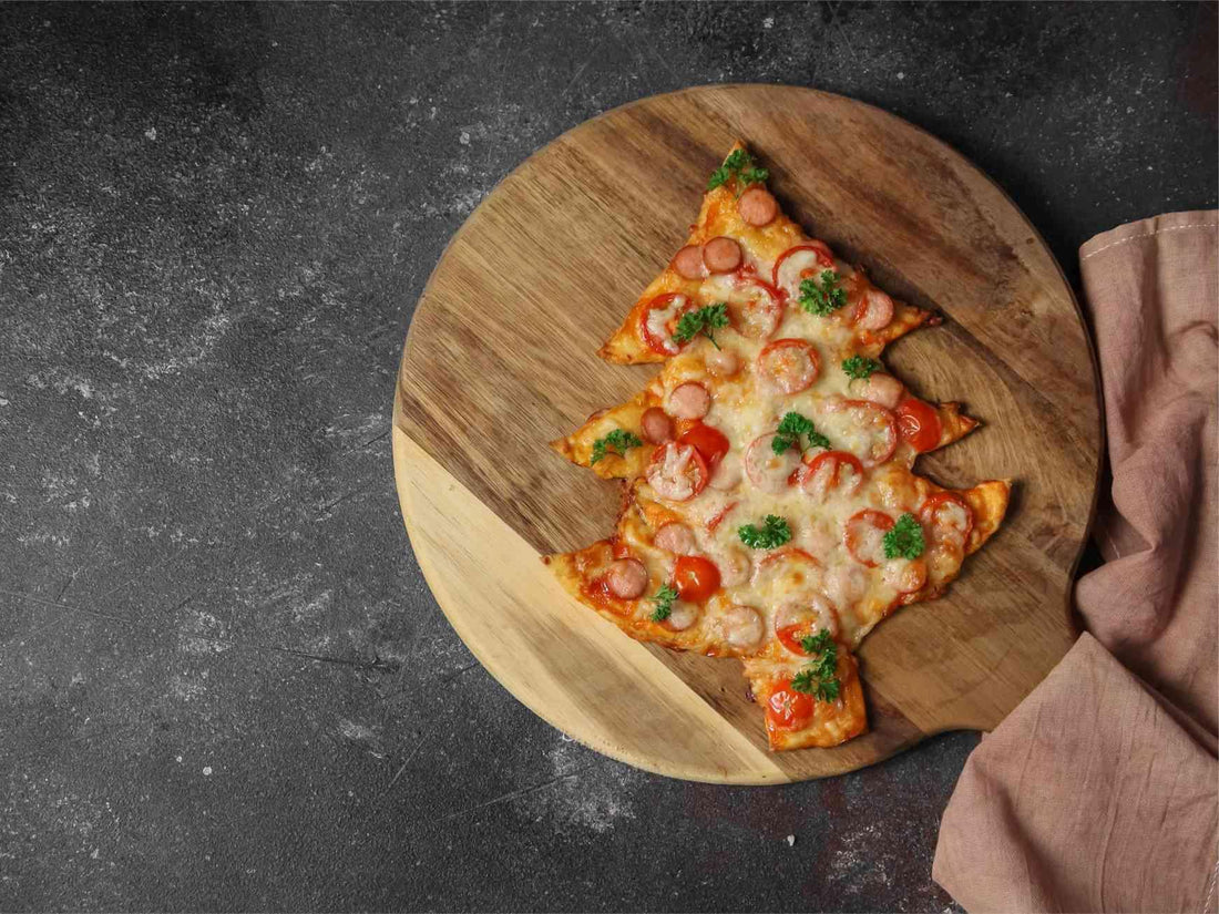 A Christmas tree-shaped pizza with cheese, pepperoni, and parsley on a wooden board against a dark surface and light brown cloth.