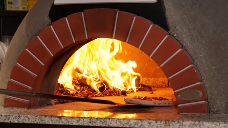 A wood-fired pizza oven with flames, cooking a pizza on stone.