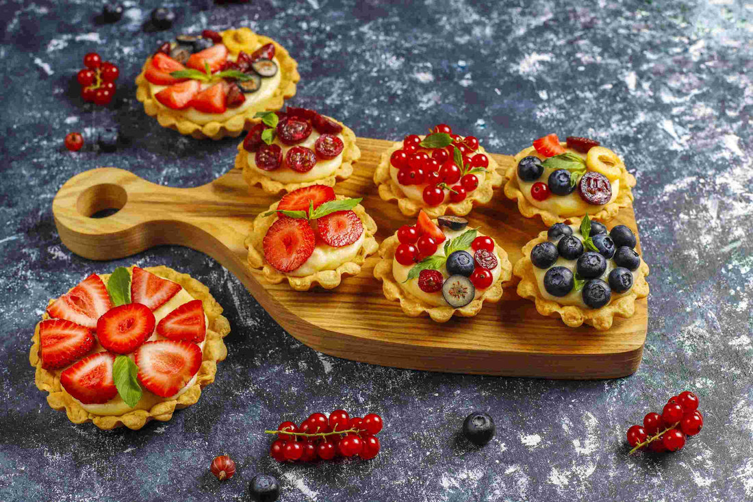 A wooden board with assorted mini fruit tarts topped with strawberries, blueberries, and red currants, surrounded by extra fruit.