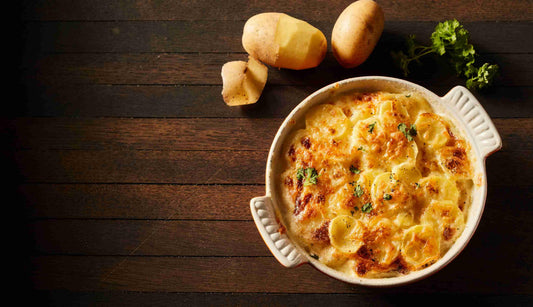 A baked potato gratin in a white ceramic dish with herbs, beside whole and peeled potatoes and parsley, on a dark wooden surface.