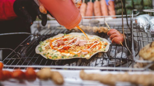 One pizza with sauce being spread, another topped with tomatoes, cheese, and mushrooms on a grill.