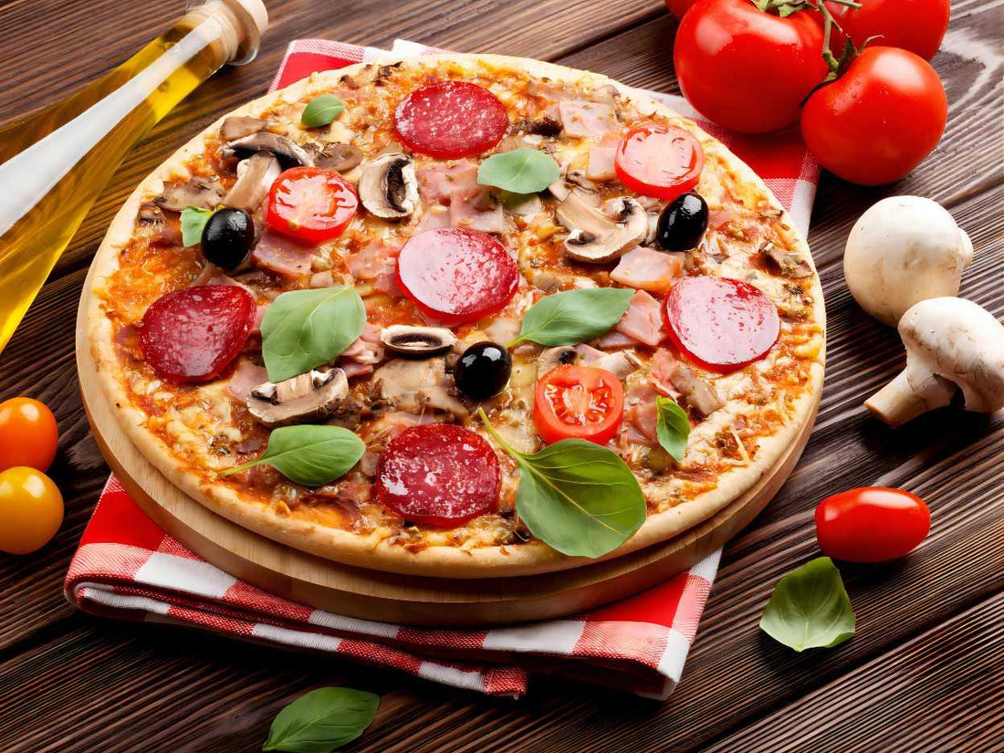 A pizza with pepperoni, tomatoes, olives, mushrooms, basil, and cheese on a wooden table with a red checkered cloth and ingredients nearby.