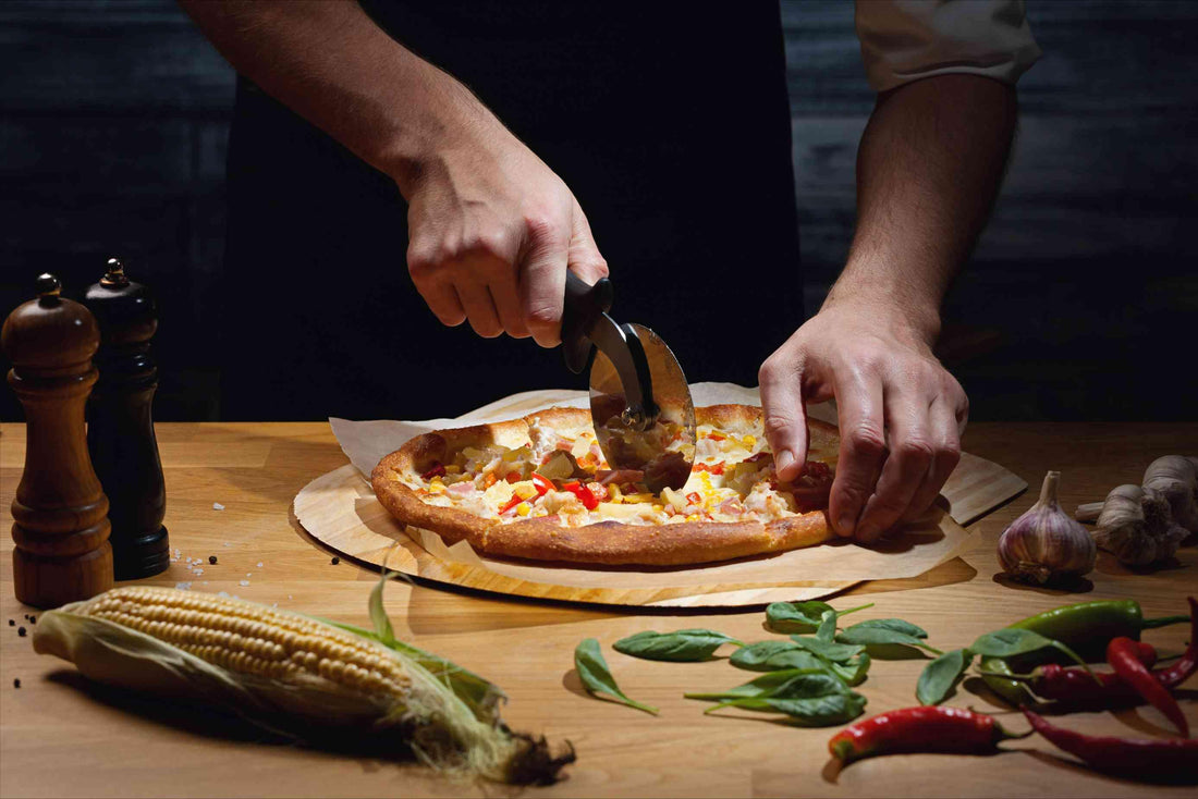 Hawaiian PizzaA person cutting a pizza on a table with spices, corn, garlic, basil leaves, and chili peppers around it. with Cauliflower Crust - Pizzello