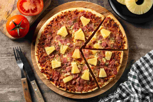 A Hawaiian pizza with ham and pineapple is sliced on a board, with utensils, a fresh tomato, and a pineapple ring nearby.