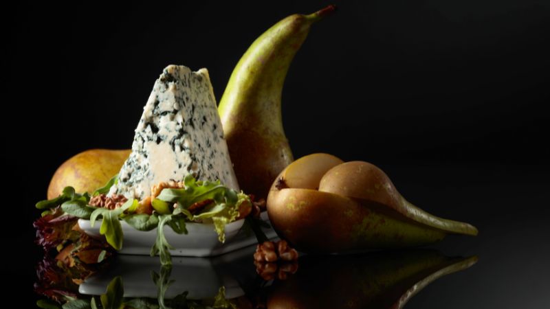 A blue cheese wedge on a white dish with pears, walnuts, and greens against a dark background.