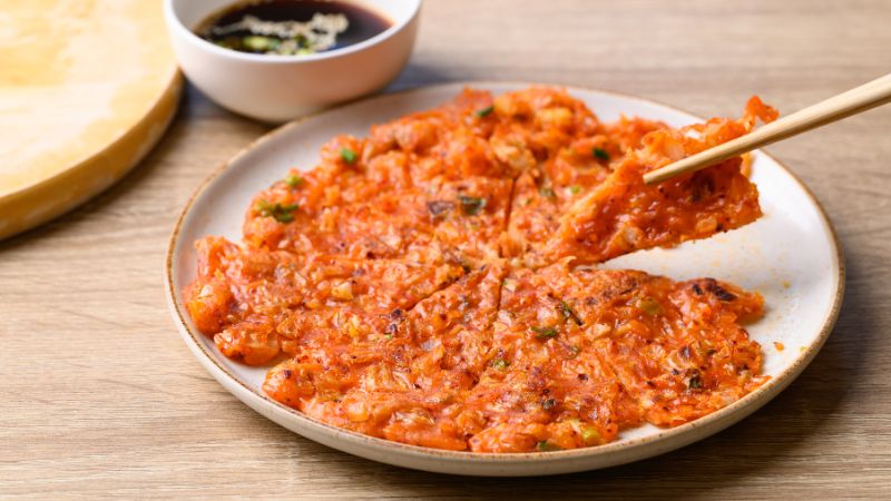 A plate of sliced kimchi pancake with one piece lifted by chopsticks, next to a small bowl of dipping sauce.