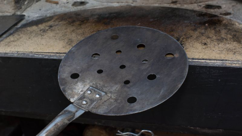 A round metal spatula with holes rests on a dark, worn surface.