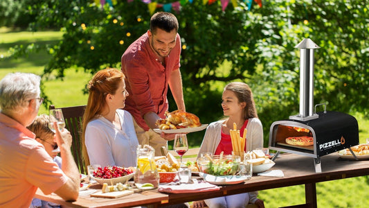 A family enjoys a meal around a table outdoors. A man serves food near a pizza oven in a bright, green garden setting.