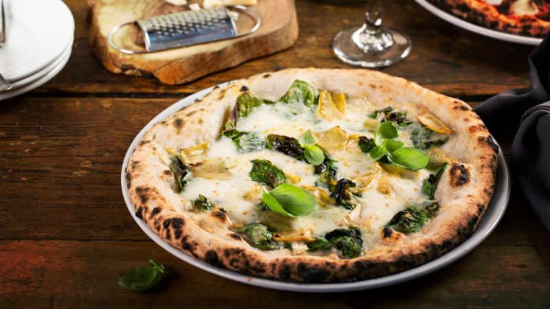 A spinach, artichoke, and basil pizza sits on a wooden table, with a cheese grater and wine glass in the background.