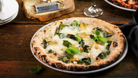 A spinach, artichoke, and basil pizza sits on a wooden table, with a cheese grater and wine glass in the background.