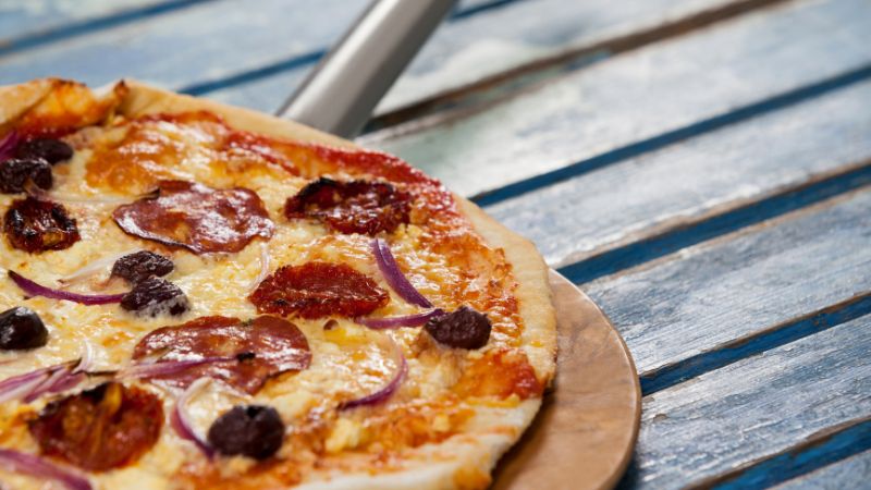 A pepperoni, red onion, and olive pizza on a wooden paddle atop a blue table.