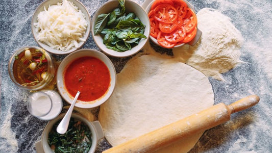 Pizza ingredients on a floured surface: dough, rolling pin, tomatoes, basil, sauce, cheese, and seasonings.