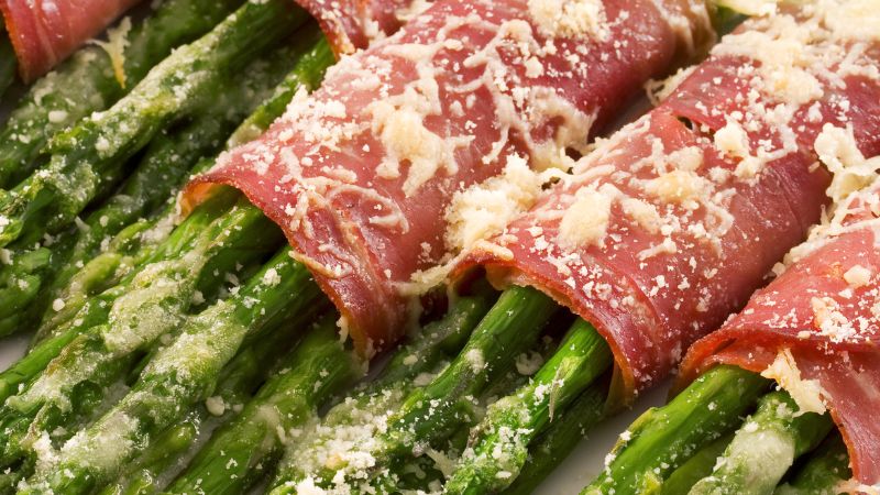 Asparagus spears wrapped in prosciutto and topped with grated cheese, arranged in rows.