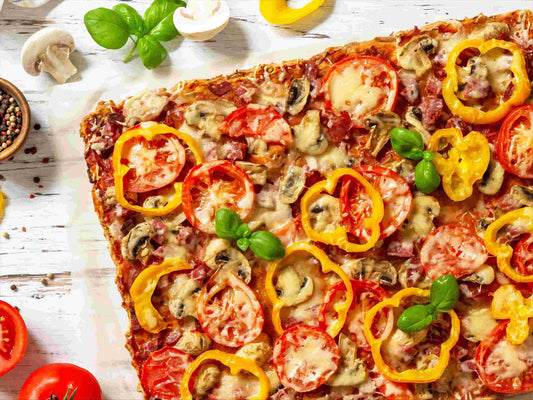 Close-up of rectangular pizza with tomatoes, yellow peppers, mushrooms, basil on a white wooden surface, scattered ingredients around