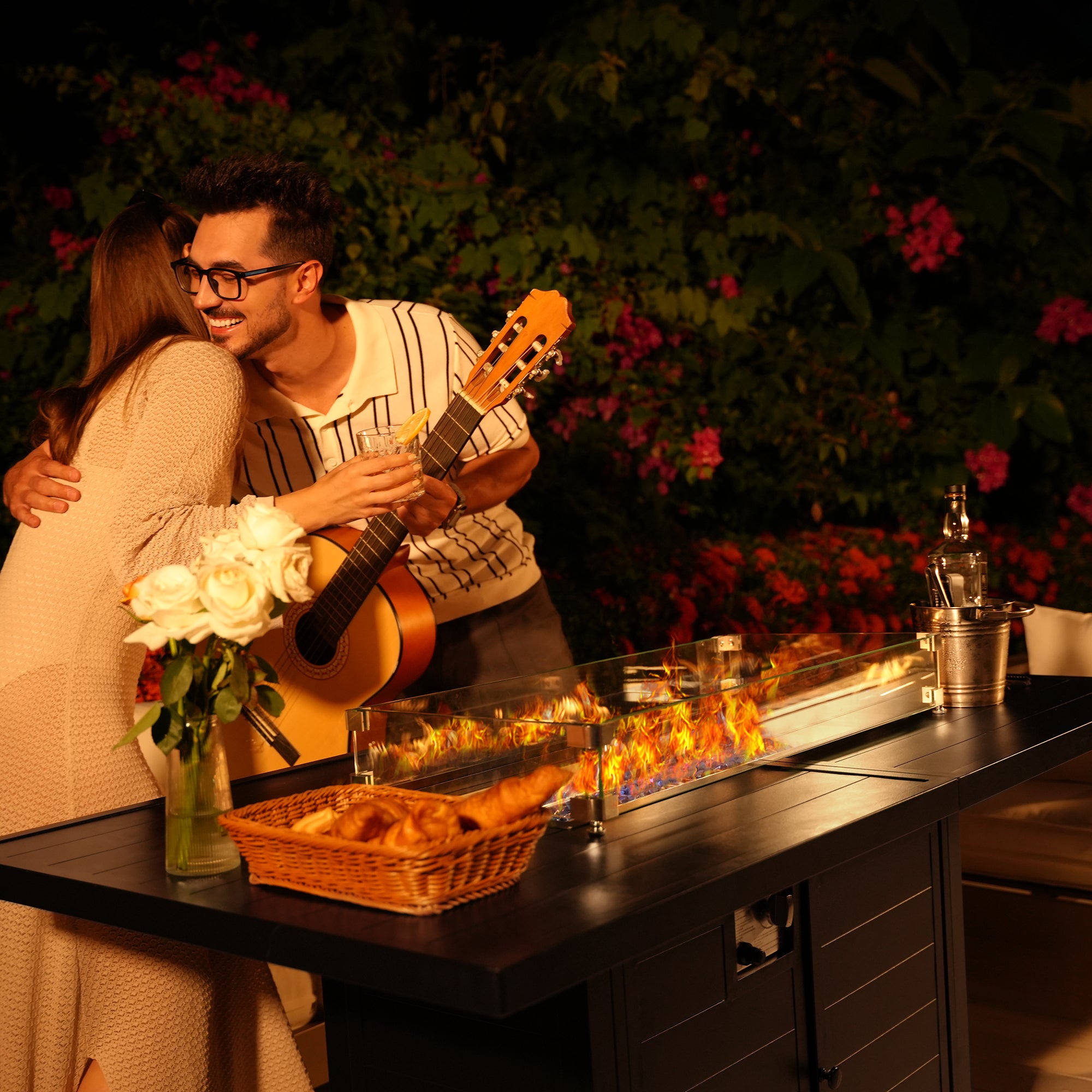 Two people embrace near a Pizzello Passione fire pit table with baguettes, flowers, a bottle, and a glass. One holds a guitar and drink.